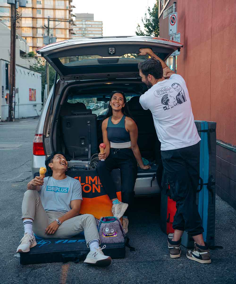 Three people gathered around the open trunk of a silver SUV in an urban street setting, with camping gear and branded bags visible. One person sits in the back of the vehicle, one stands opening the trunk, and one sits outside smiling with an ice cream cone.