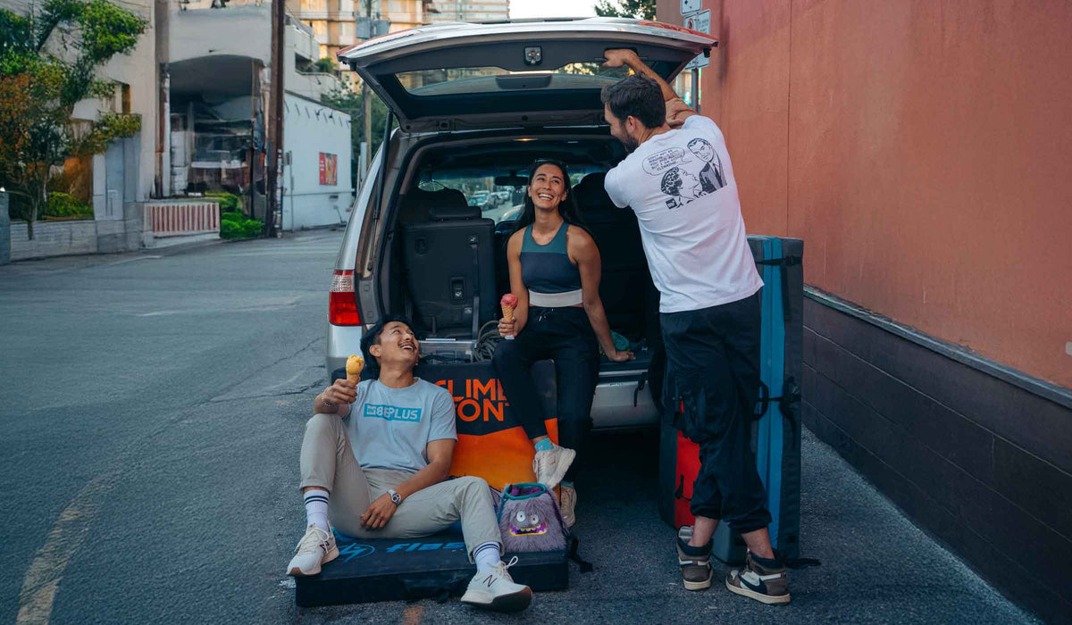 Three people gathered around the open trunk of a white SUV in an outdoor street setting, with one person sitting on the ground holding an ice cream cone, another standing in the back of the vehicle, and a third person adjusting the open trunk. A blue and black travel luggage bag is visible on the right side.
